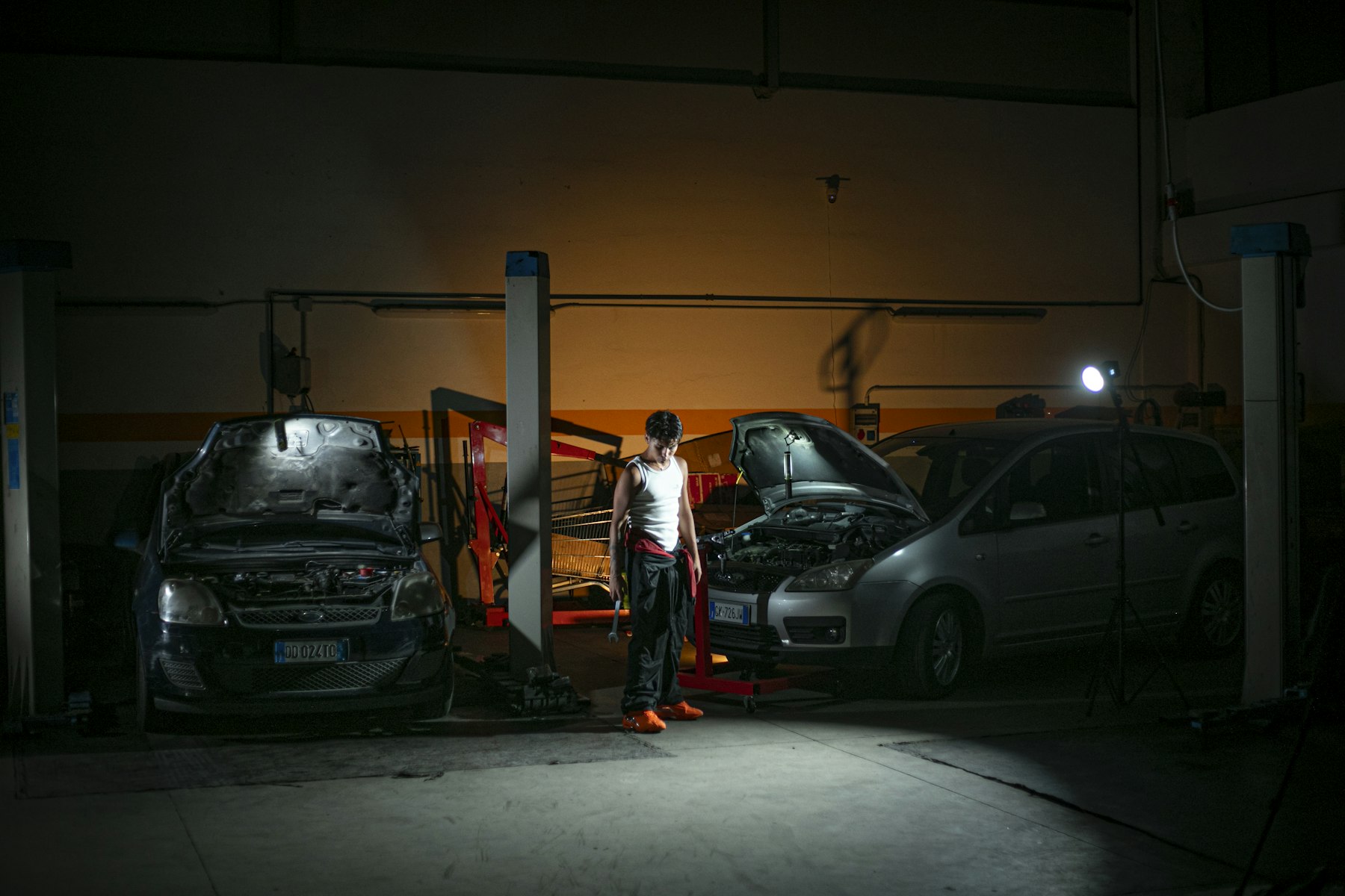 Workshop service bays at night with two cars under work lights, bonnets up between the hoists