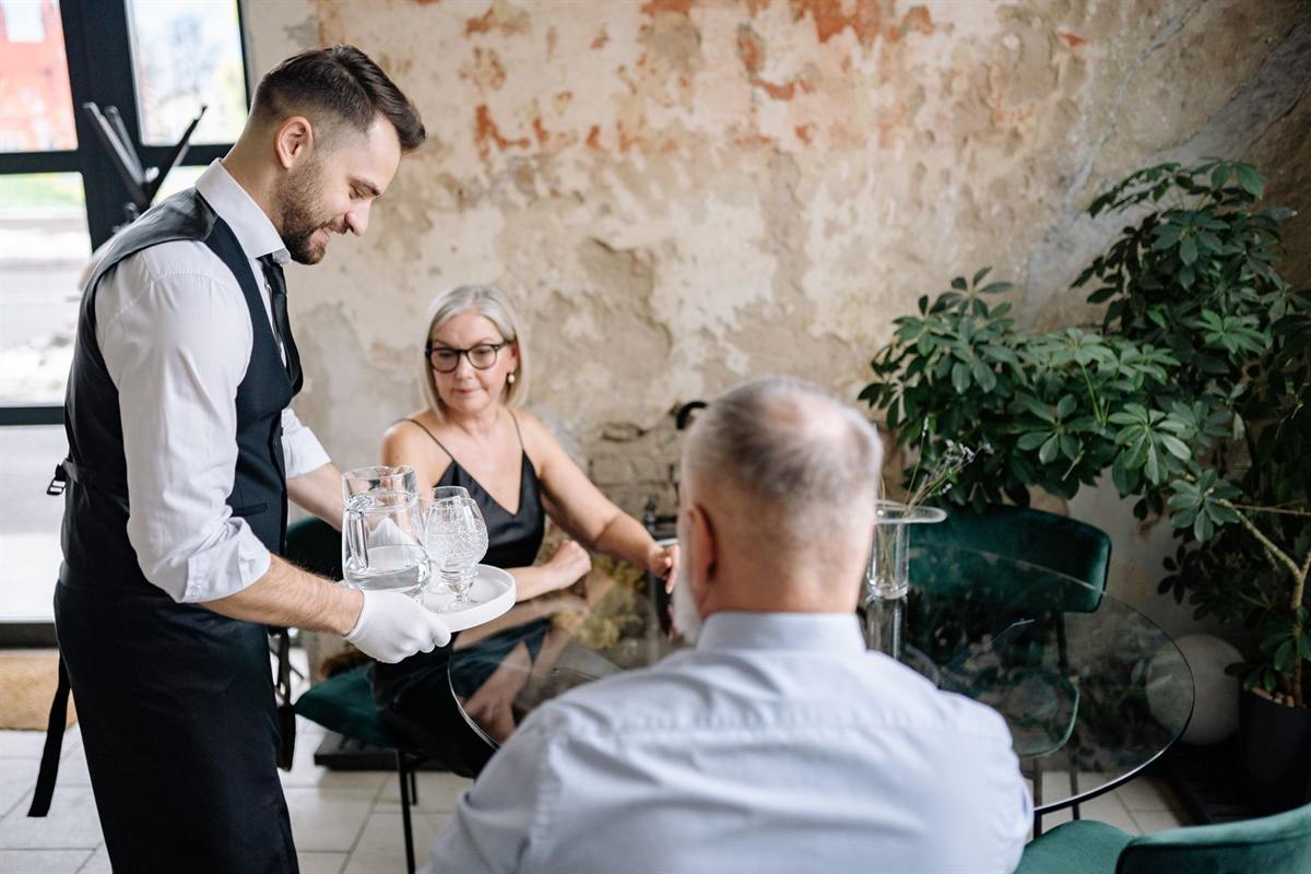 Waiter in a black apron serving water to two seated diners in a rustic restaurant